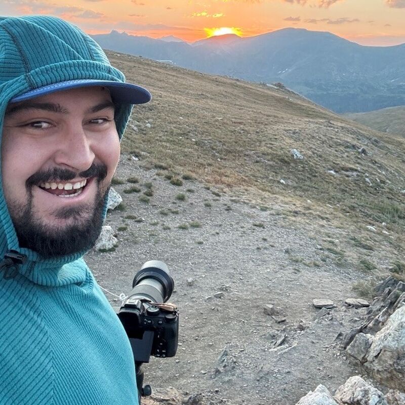 A man with a beard and a blue hooded sweatshirt is taking a selfie on a mountain. He is holding a camera with a large lens. The sun is setting in the background, casting a warm glow over the landscape. The terrain appears rocky and grassy, suggesting a high-altitude environment. The man is smiling, indicating he is enjoying the scenery.